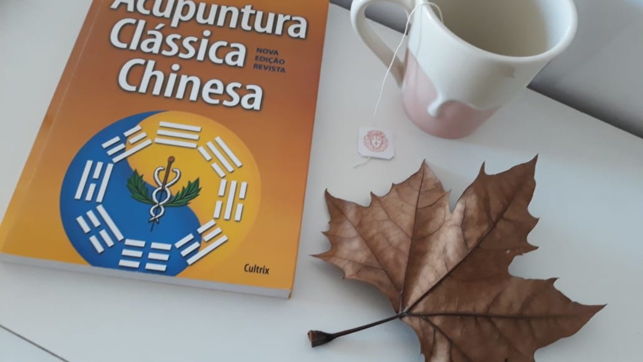 Foto de livro de acupuntura, xícara e folha seca numa mesa